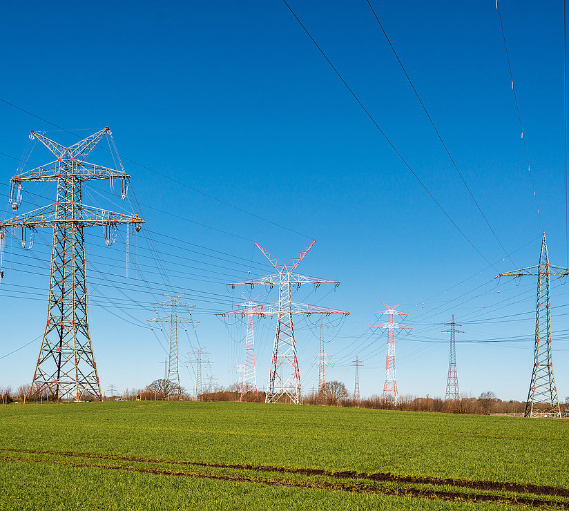 Mehrere Strommasten und Stromleitungen erstrecken sich über eine grüne Wiese unter einem klaren blauen Himmel und transportieren Strom über weite Strecken.
