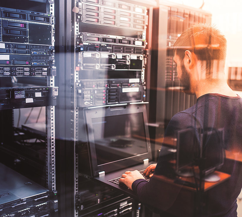 Ein Mann steht in einem Serverraum und arbeitet an einem Laptop, der an ein Server-Rack voller Geräte und Kabel angeschlossen ist. Durch eine Glaswand wird die Szene von einem warmen Licht erhellt.